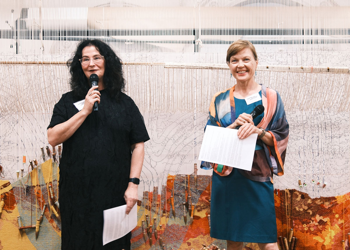 Two women stand in front of a loom, piles of colourful yarn in front of them