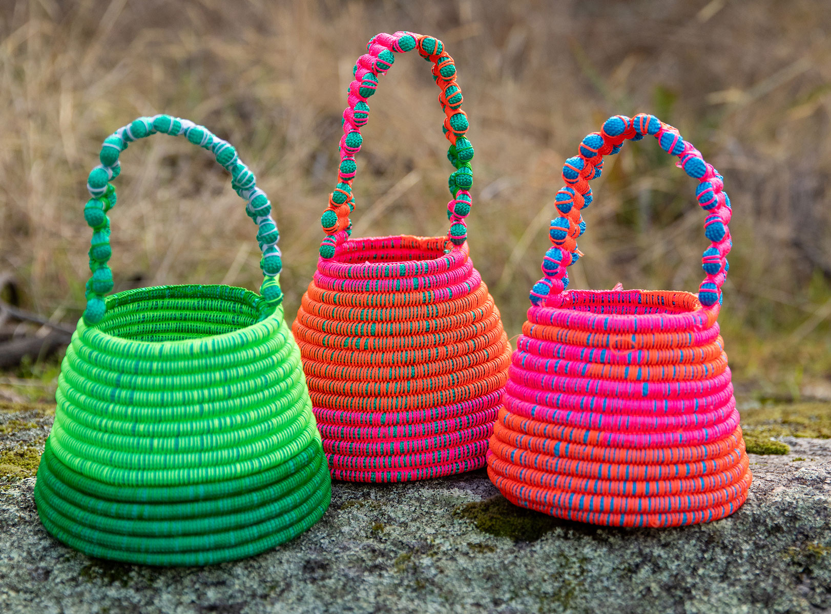 3 brightly coloured a coiled vessel sit on a rock with tall grass in the background