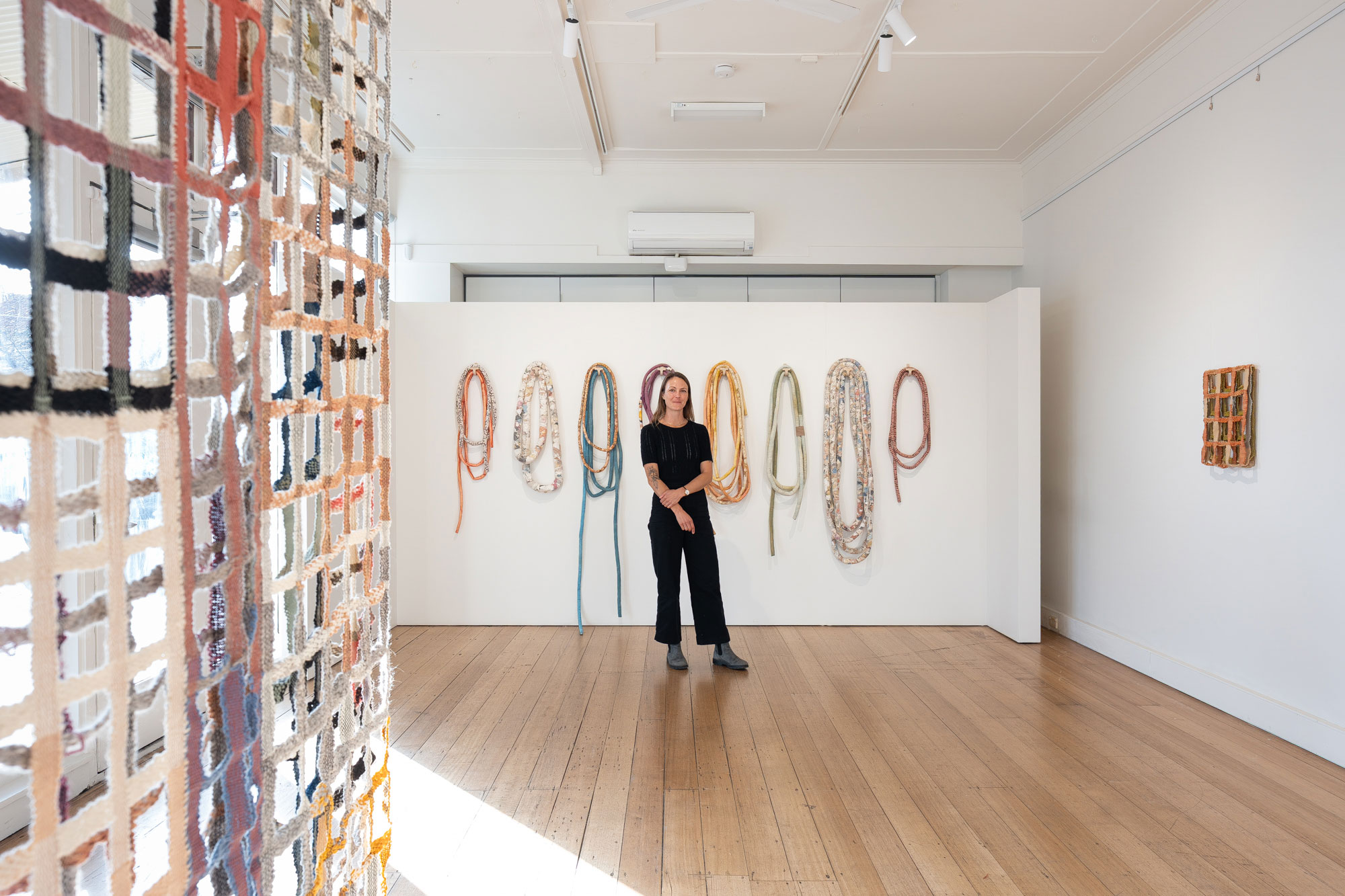 A woman stands in a gallery space amongst her textile artworks, some artworks resemble nets or trellises, while others look like long sausages looped around. The colours are muted.