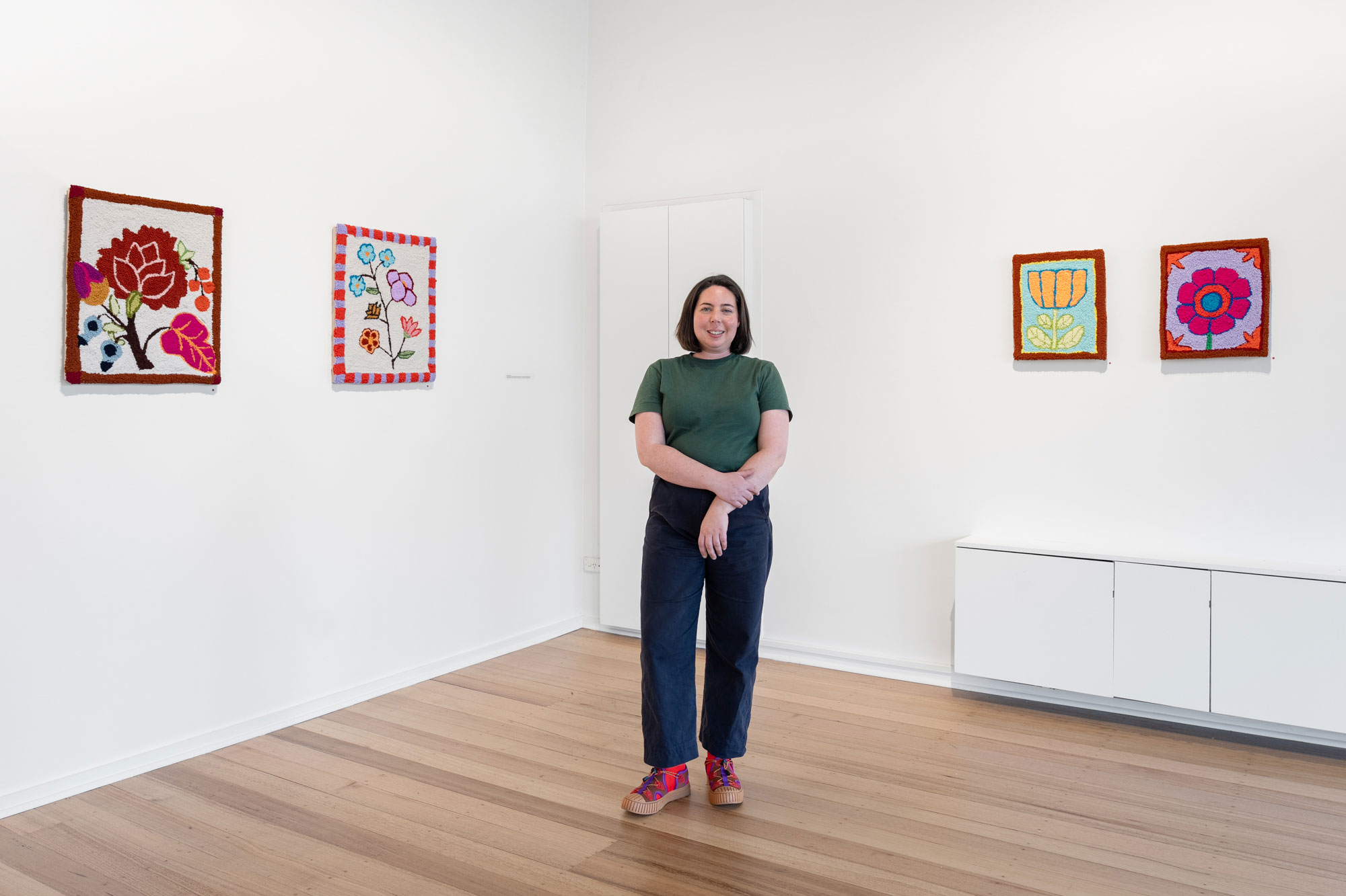 A woman stands in a gallery space amongst her brightly coloured artworks